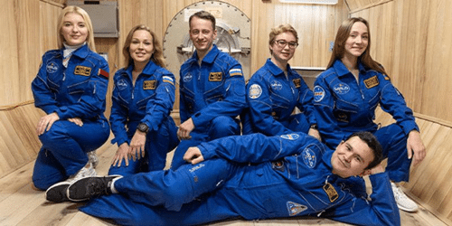six people in blue space suits in front of a wooden wall with a hatch behind them one man laying down in front of the others who are squatting in a paint me like your french girls pose m.png