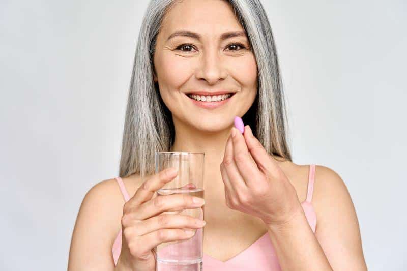 grey haired woman in pink tank top taking a pink pill holding a glass of water m.jpg