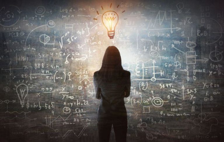 person with long hair in business suit facing away from the camera towards blackboard with chalk equations on illustrated lightbulb above their head m.jpg