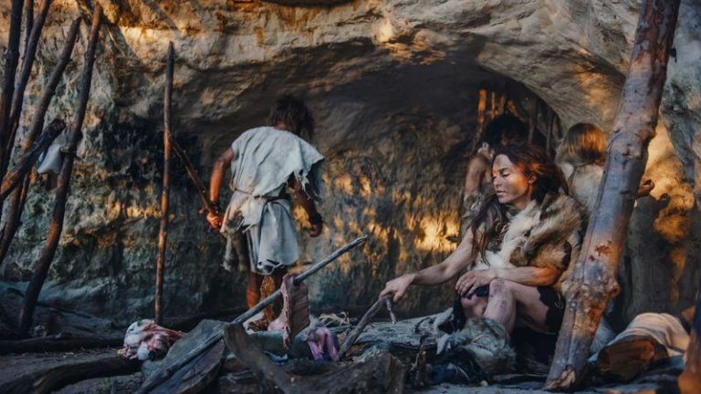 prehistoric looking woman poking a fire with a stick sat by a cave other individuals in the background m.jpg