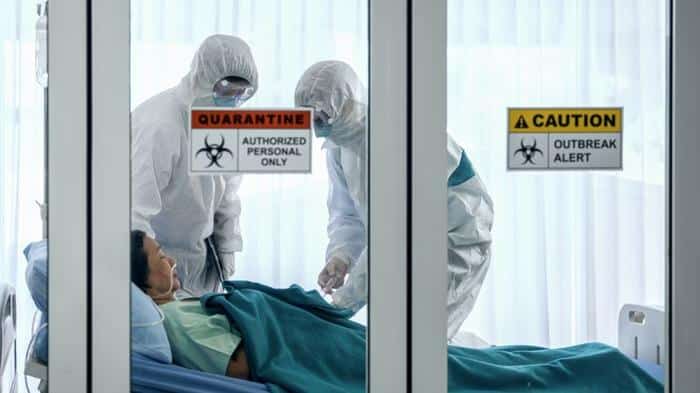 two people in white protective suits and masks standing over a person in a hospital bed with tubes in their nose the glass door has quarantine warning signs on it m.jpg