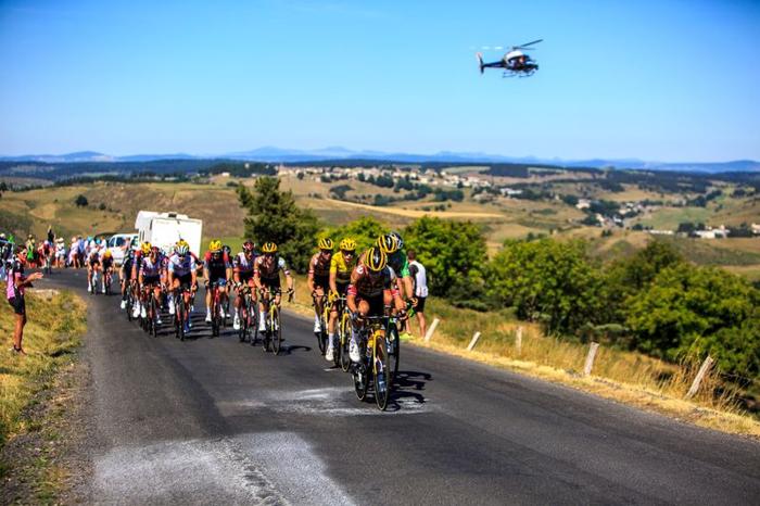 large group of cyclists during the tour de france on a road lines with greenery a helicopter in the blue sky behind them m.jpg