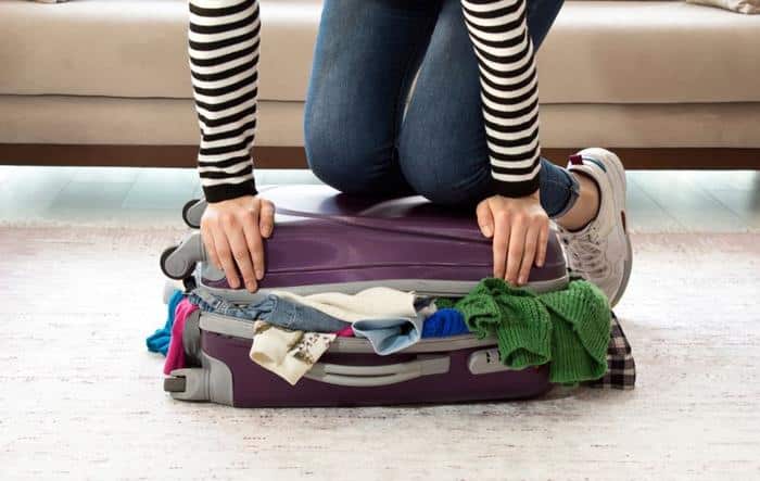 person wearing jeans and striped top kneeling on purple overfilled suitcase that will not shut m.jpg